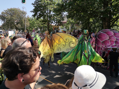 Farbfoto von drei als Schmetterling verkleideten Stelzenläufern auf dem Umzug vom Karneval der Kulturen auf der Straße Hasenheide in Berlin am 28. Mai 2023. Fotograf: Erwin Thomasius.